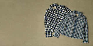Two cropped button-up plaid shirts laid flat on a textured beige background. One shirt has a navy and white plaid pattern, and the other has a blue and white plaid pattern.