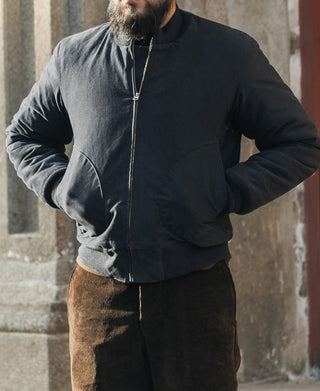 Person wearing a dark jacket and brown pants standing against a stone wall.